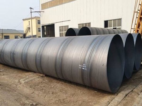 Se observan Tubería de Diámetros Mayores de acero, alineadas en una zona exterior cerca de un edificio industrial. Las tuberías tienen un acabado liso y están dispuestas de manera ordenada sobre el suelo.