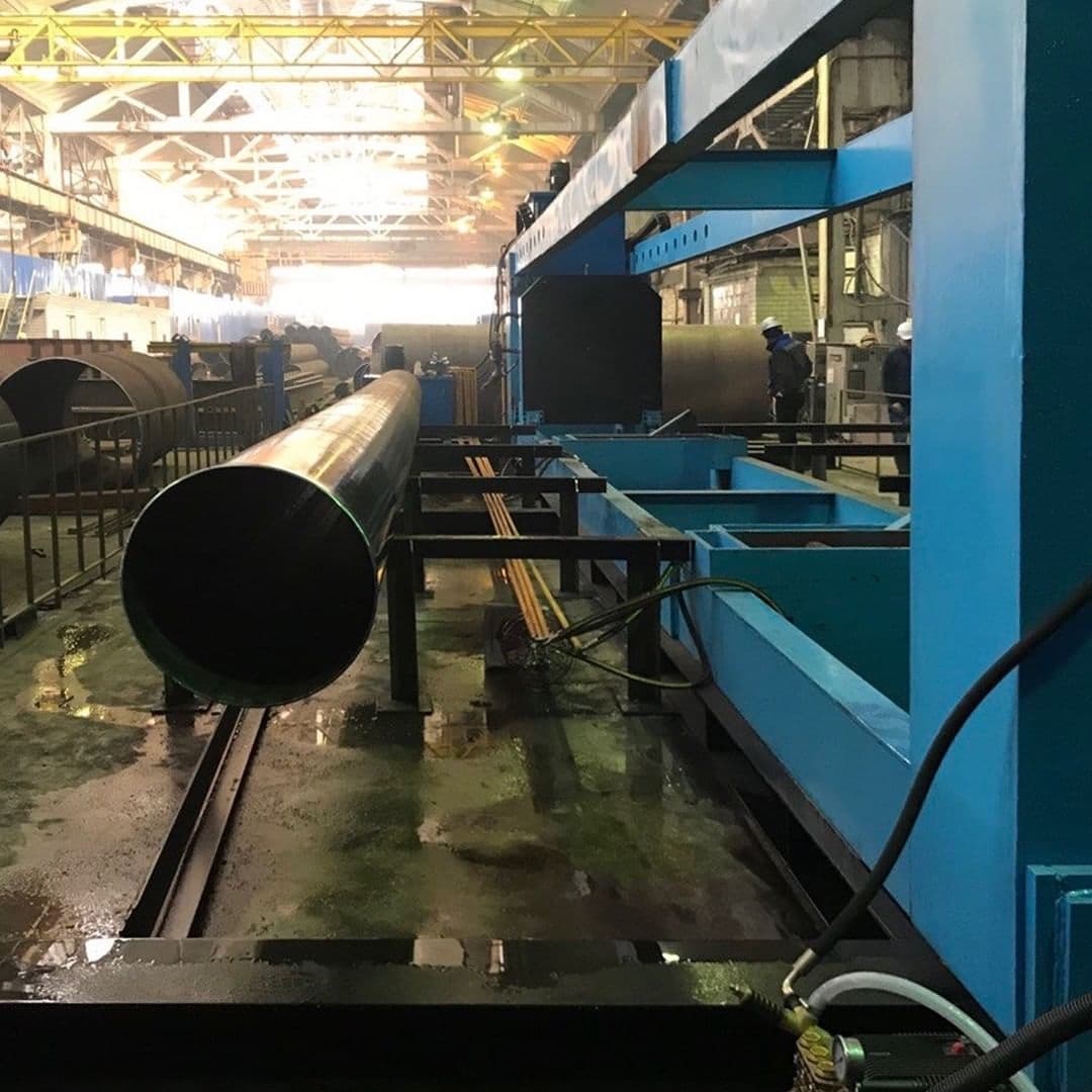 Instalación industrial con maquinaria para el procesamiento de Tubería de Diámetros Mayores. Se observa una tubería larga en primer plano y equipos de trabajo en segundo plano, bajo una iluminación brillante.