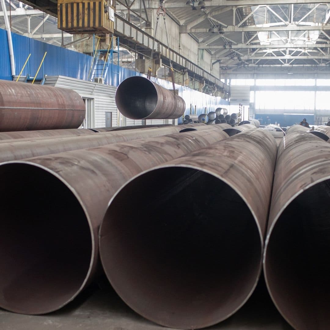 Tubería de Diámetros Mayores apiladas en un entorno industrial, con una grúa en el fondo y una luz natural que entra por las grandes ventanas.