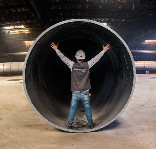 Un hombre se encuentra de pie dentro de una tubería de Diámetros Mayores, levantando los brazos hacia los lados, en un entorno industrial con iluminación tenue.