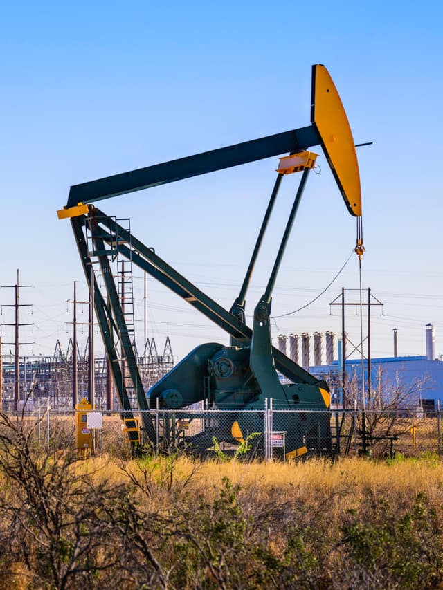 Bomba de extracción de petróleo en un campo industrial rodeada de vegetación seca y torres eléctricas, con un cielo despejado de fondo. Ideal para la producción de Tubería de Diámetros Mayores para la industria petrolera.